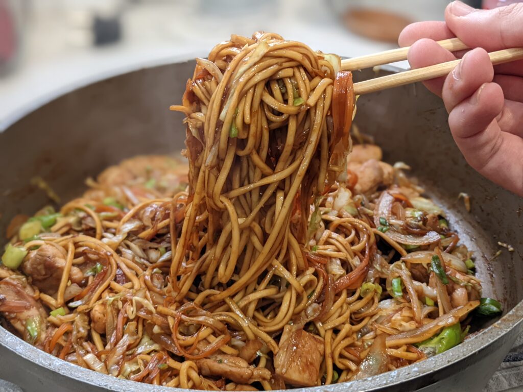 Dark egg noodle chicken stir fry being lifted with chopsticks from a wok