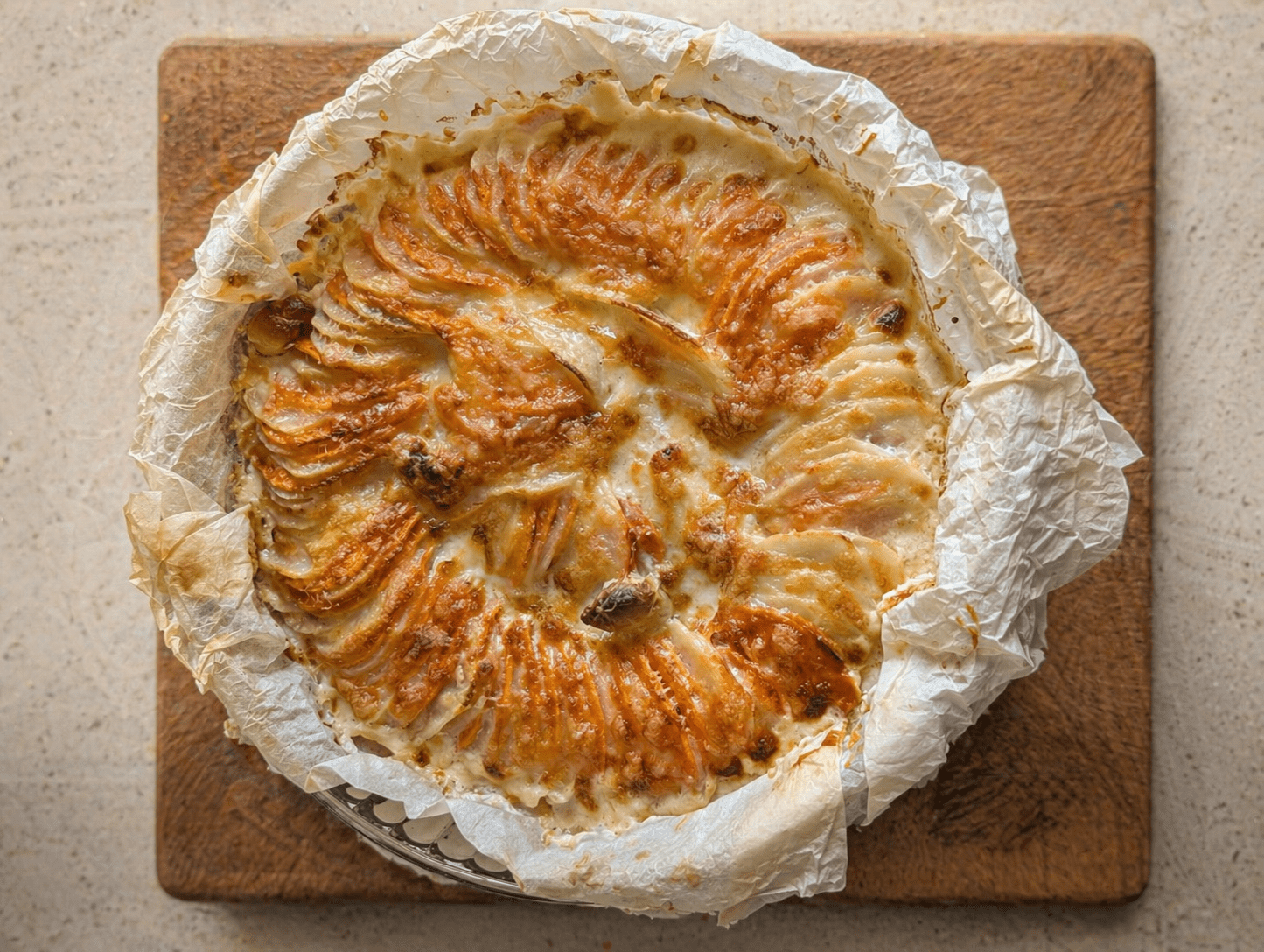 Two-tone scalloped potatoes baked golden in a round dish on a wooden board, overhead view