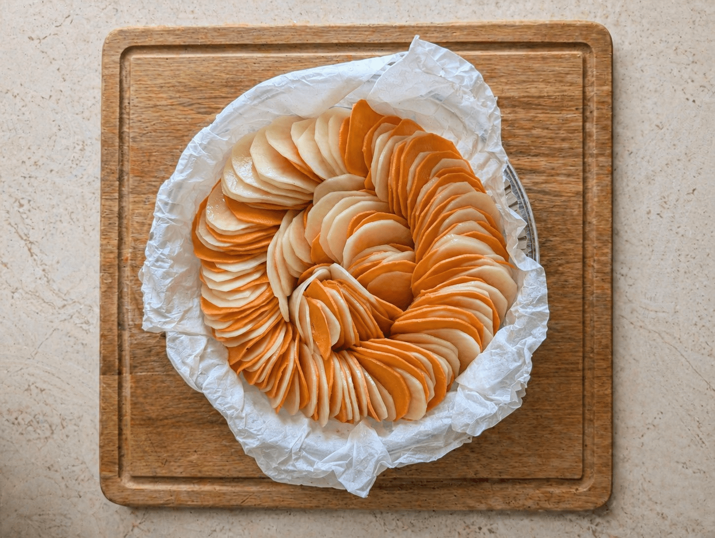Raw two-tone scalloped potatoes arranged in a spiral in a round parchment-lined pan, alternating orange and white slices