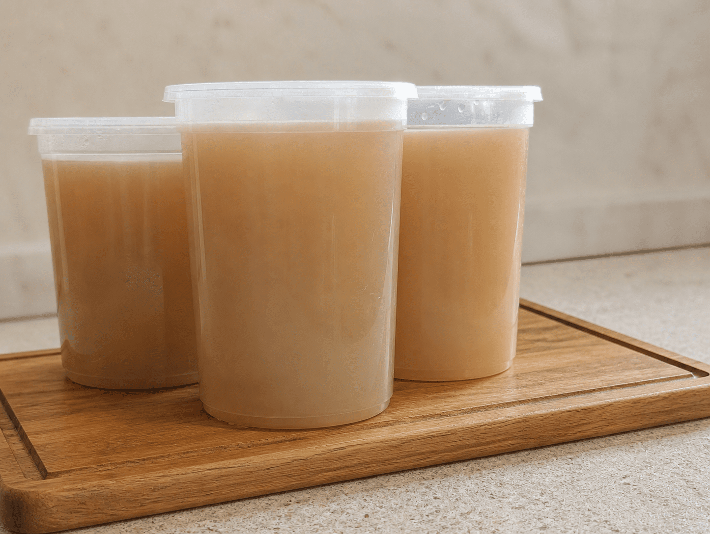 Three plastic containers full of golden homemade chicken bone broth on a wooden board