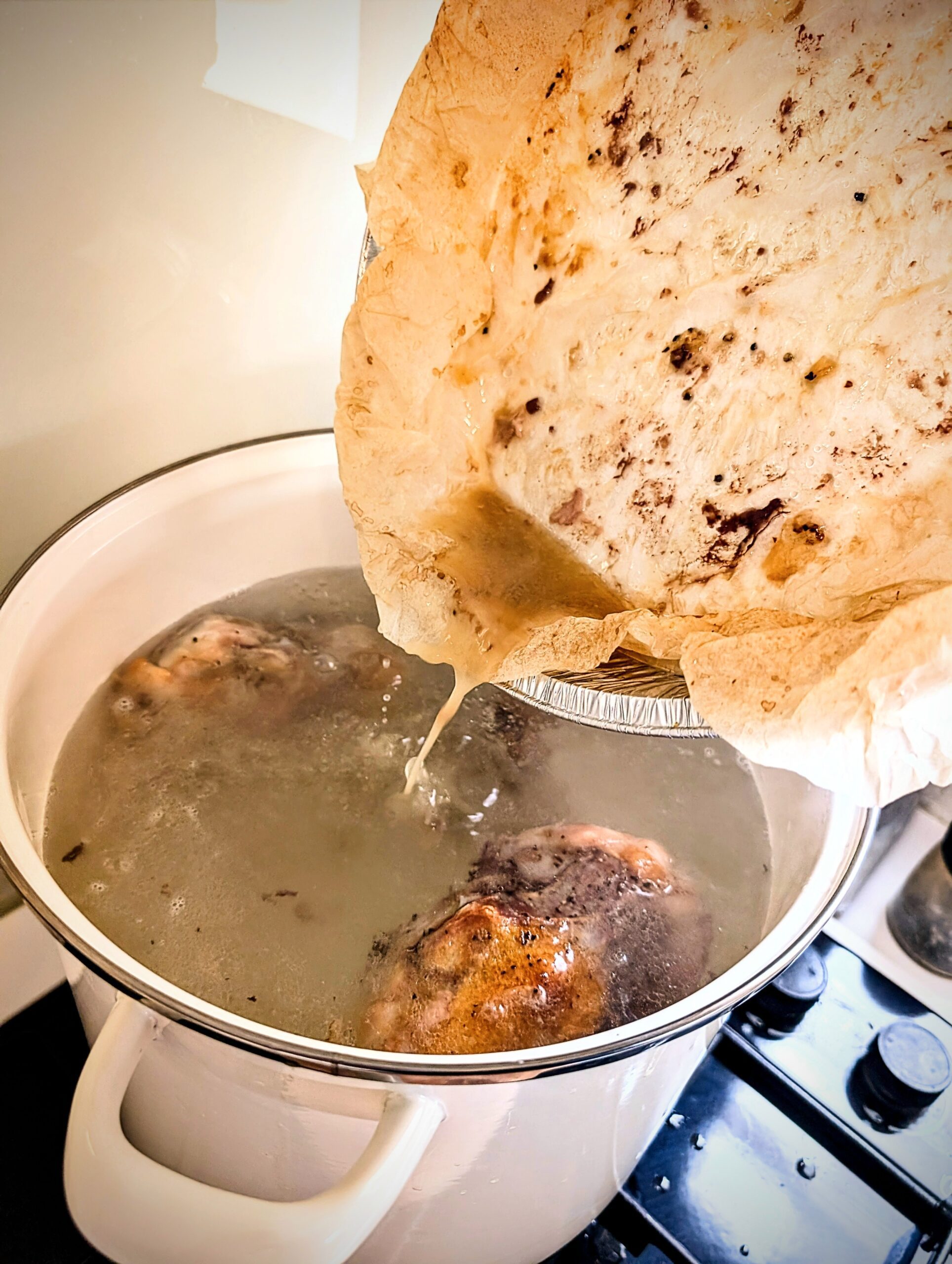 Pouring rendered bone juices from the roasting tray into a large pot of simmering water with chicken bones