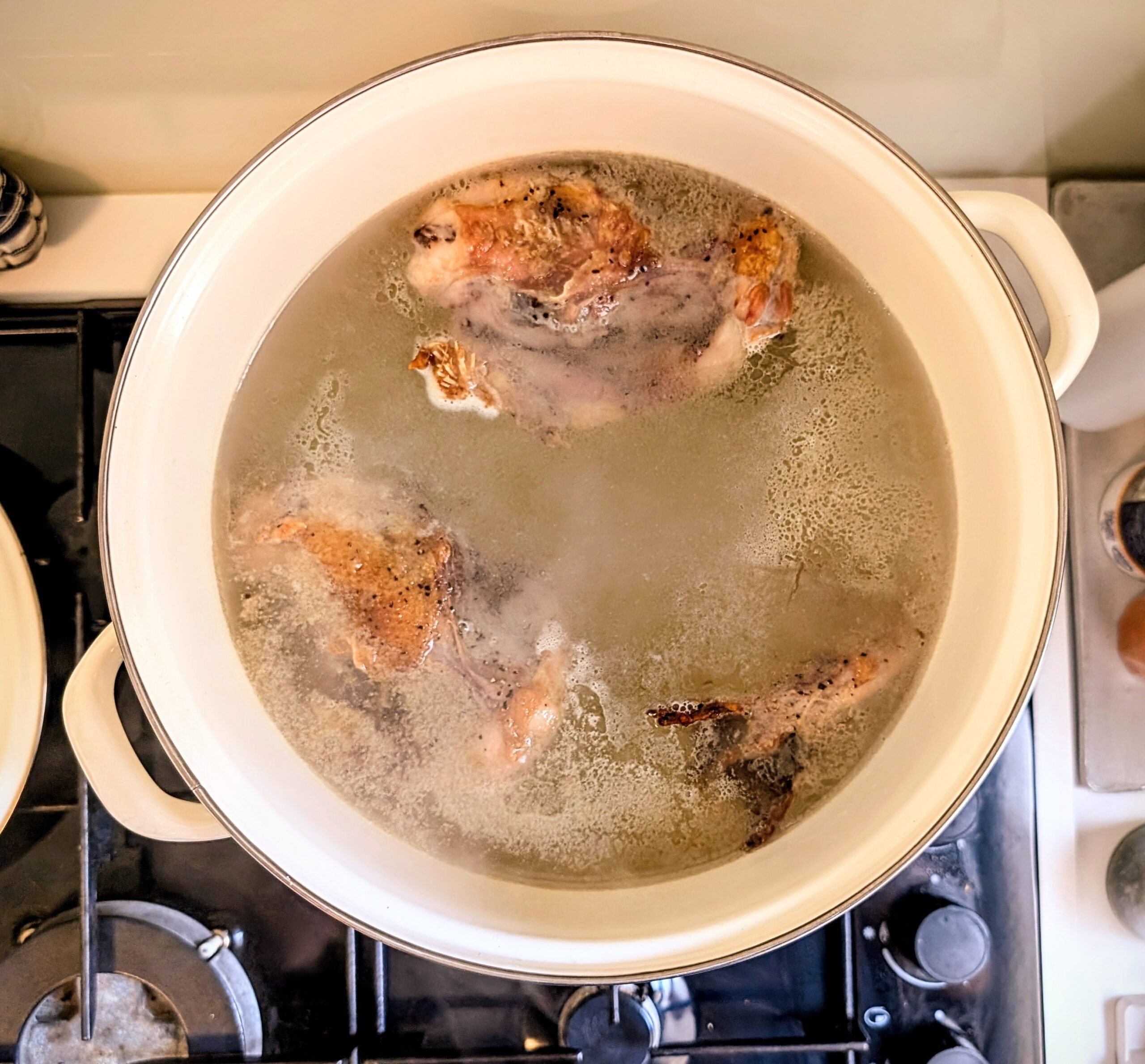Overhead view of roasted chicken bones simmering in a large white pot of clear broth on the stove