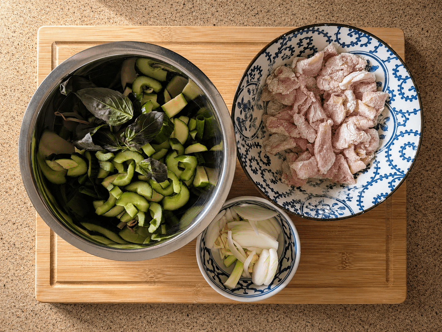 Mise en place for green curry noodle stir fry showing prep bowls: sliced cucumber and zucchini with basil, raw chicken in a blue bowl, and a small bowl of sliced onion and garlic