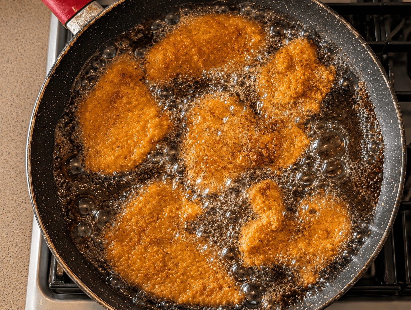 Several Israeli chicken schnitzel cutlets shallow-frying in bubbling hot oil in a large black pan, turning golden brown on the edges
