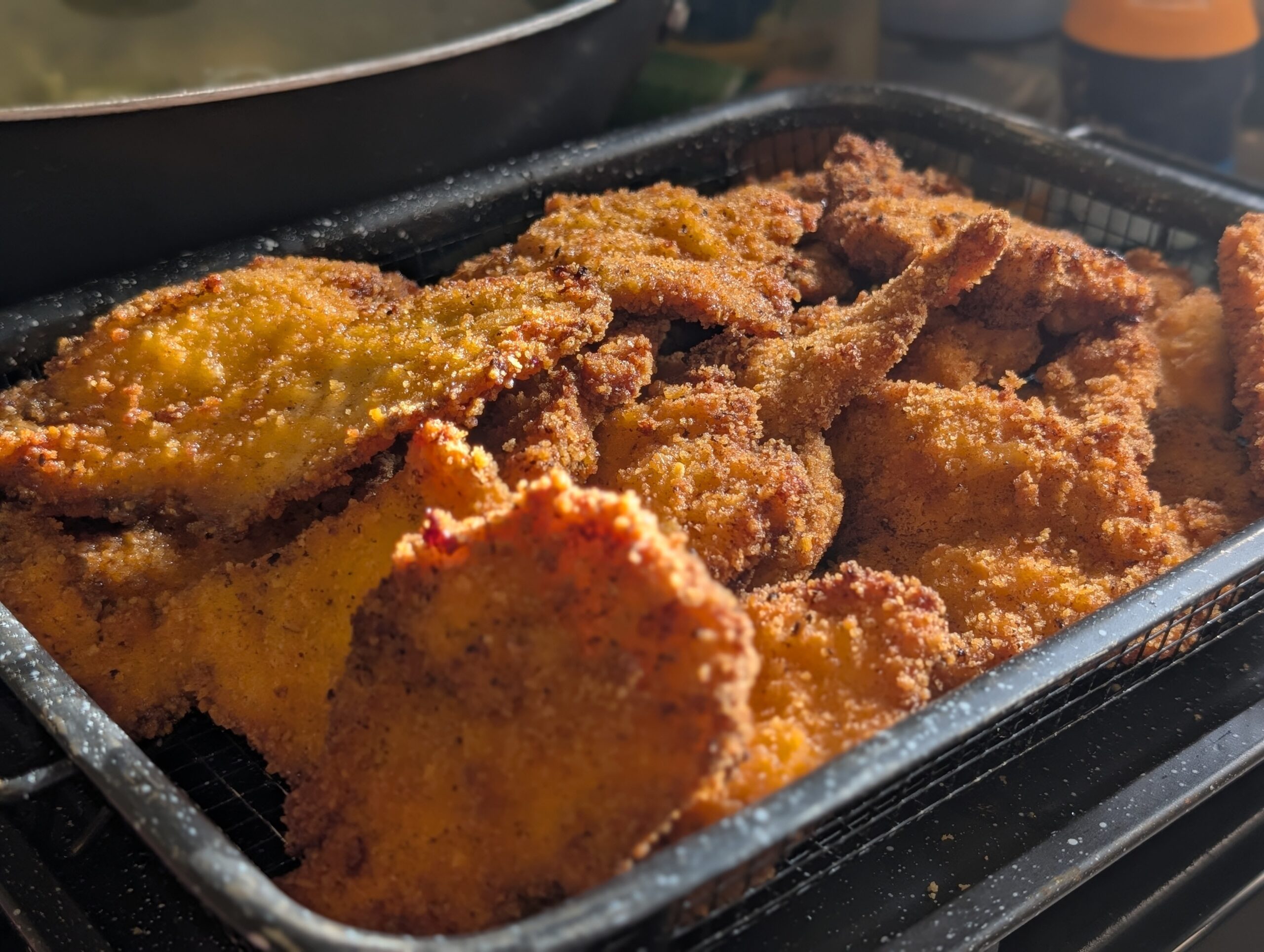 Pile of crispy golden Israeli chicken schnitzel on a wire cooling rack inside a black baking tray, fresh from the fryer