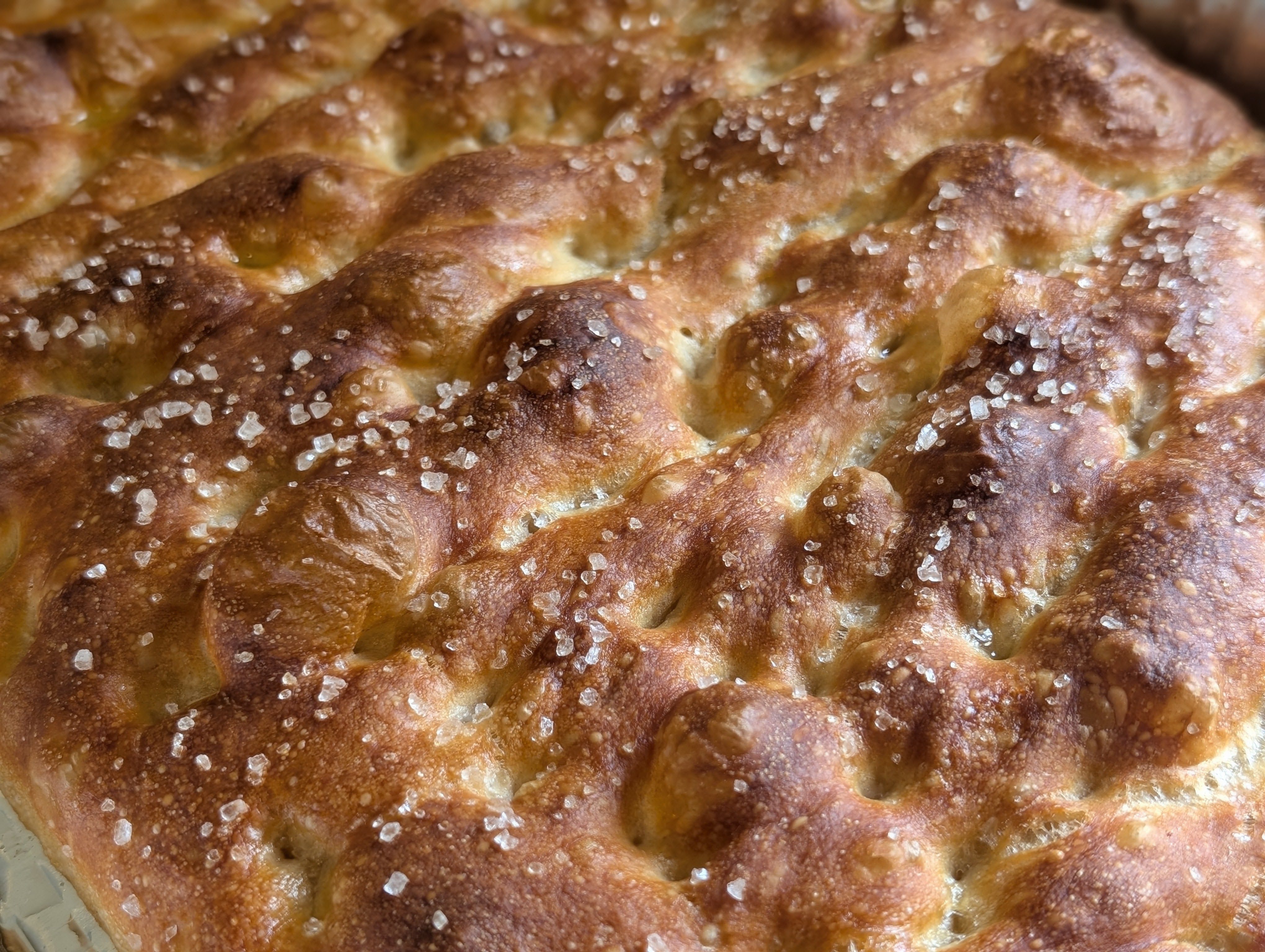 Close-up of baked no knead focaccia top showing deep golden brown crust, deep dimples, and flaky salt crystals scattered across the blistered surface