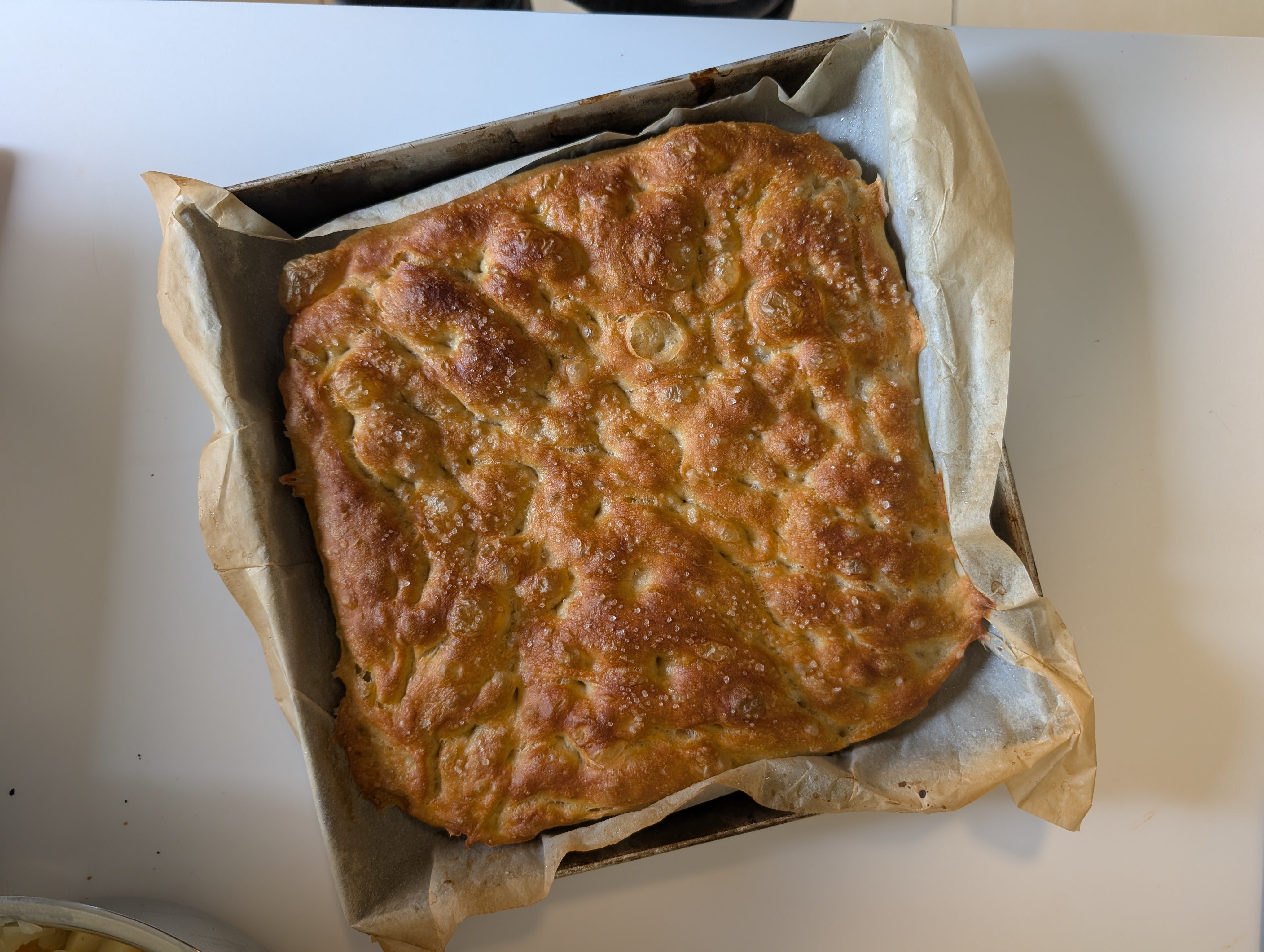 Top-down view of baked no knead focaccia in a parchment-lined baking tray on a white countertop, deeply golden across the surface