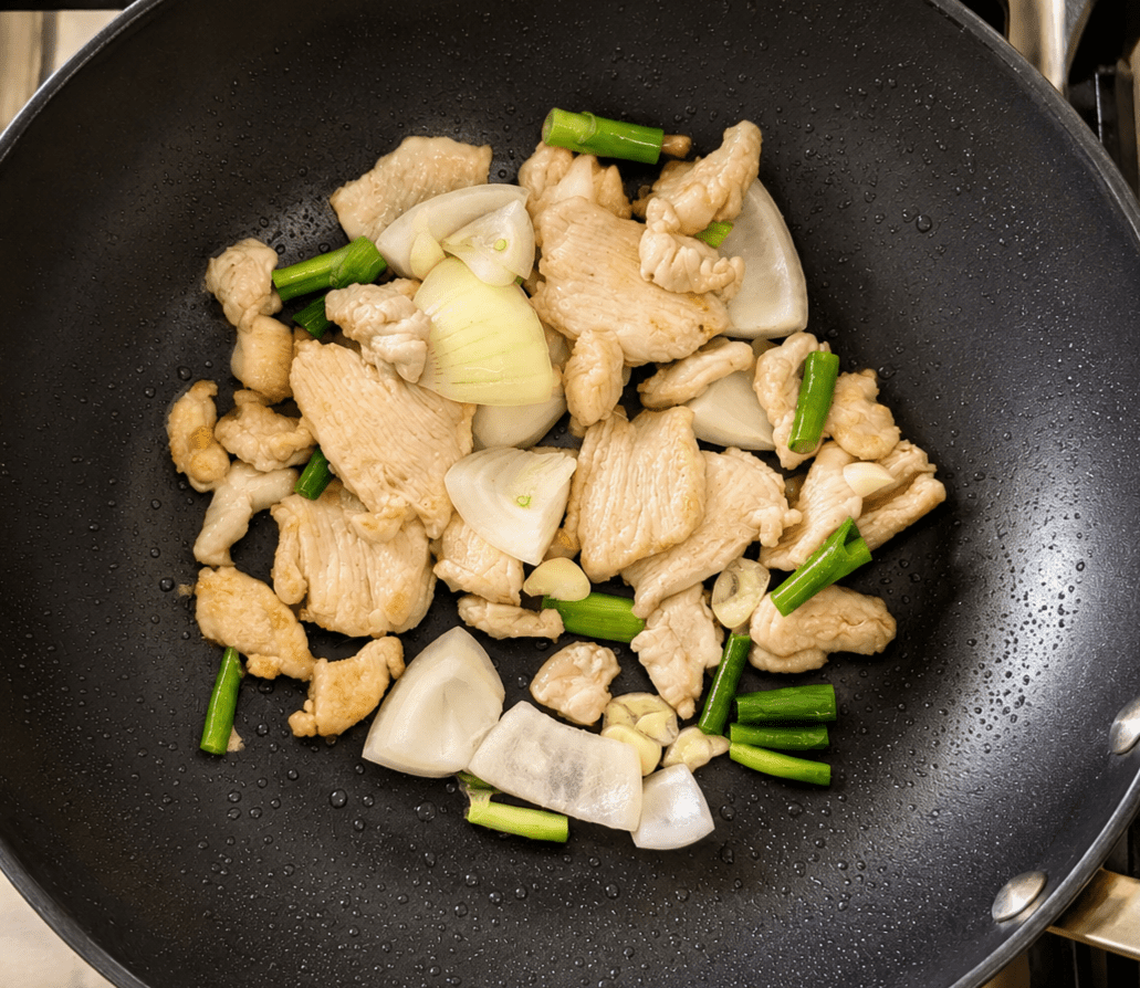 Sliced chicken pieces stir-frying in a wok with chunks of white onion, garlic and green onion batons