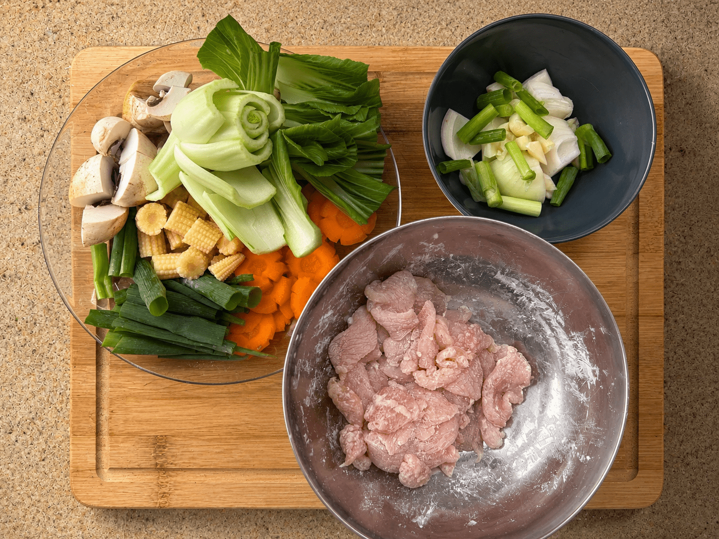 Mise en place for oyster sauce chicken stir fry showing prep bowls: chopped vegetables in a glass bowl, sliced onion and green onions in a small dark bowl, and corn-flour-coated chicken in a metal bowl