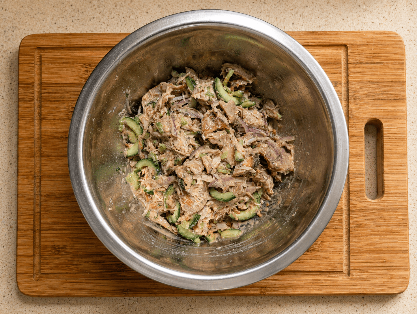 Finished leftover rotisserie chicken salad in a stainless steel bowl on a wooden board, with visible pieces of cucumber, red onion and dill