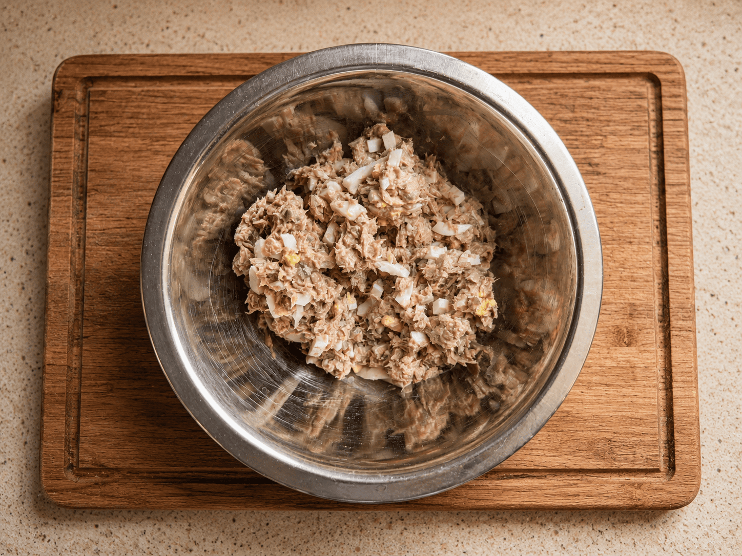 Finished tuna salad mixed in a metal bowl on a wooden board, showing visible chunks of egg and pickle