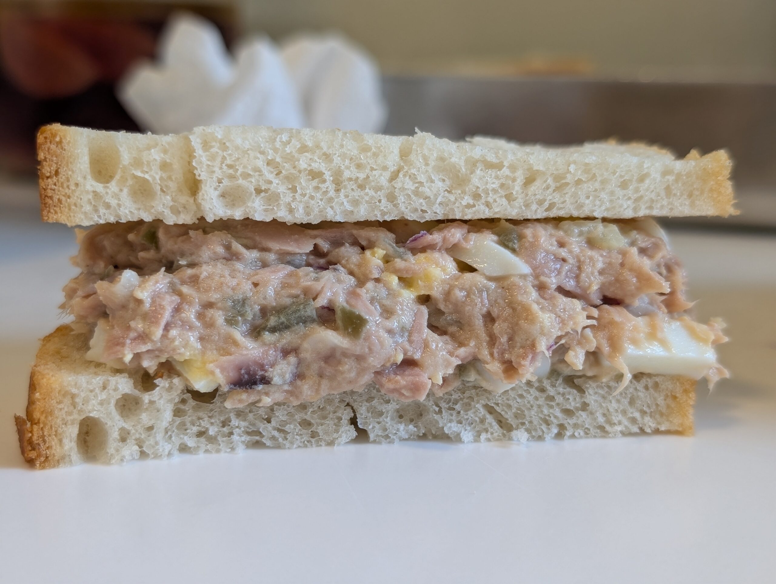 Side cross-section of a tuna salad sandwich on white bread showing the textured filling with visible egg pieces