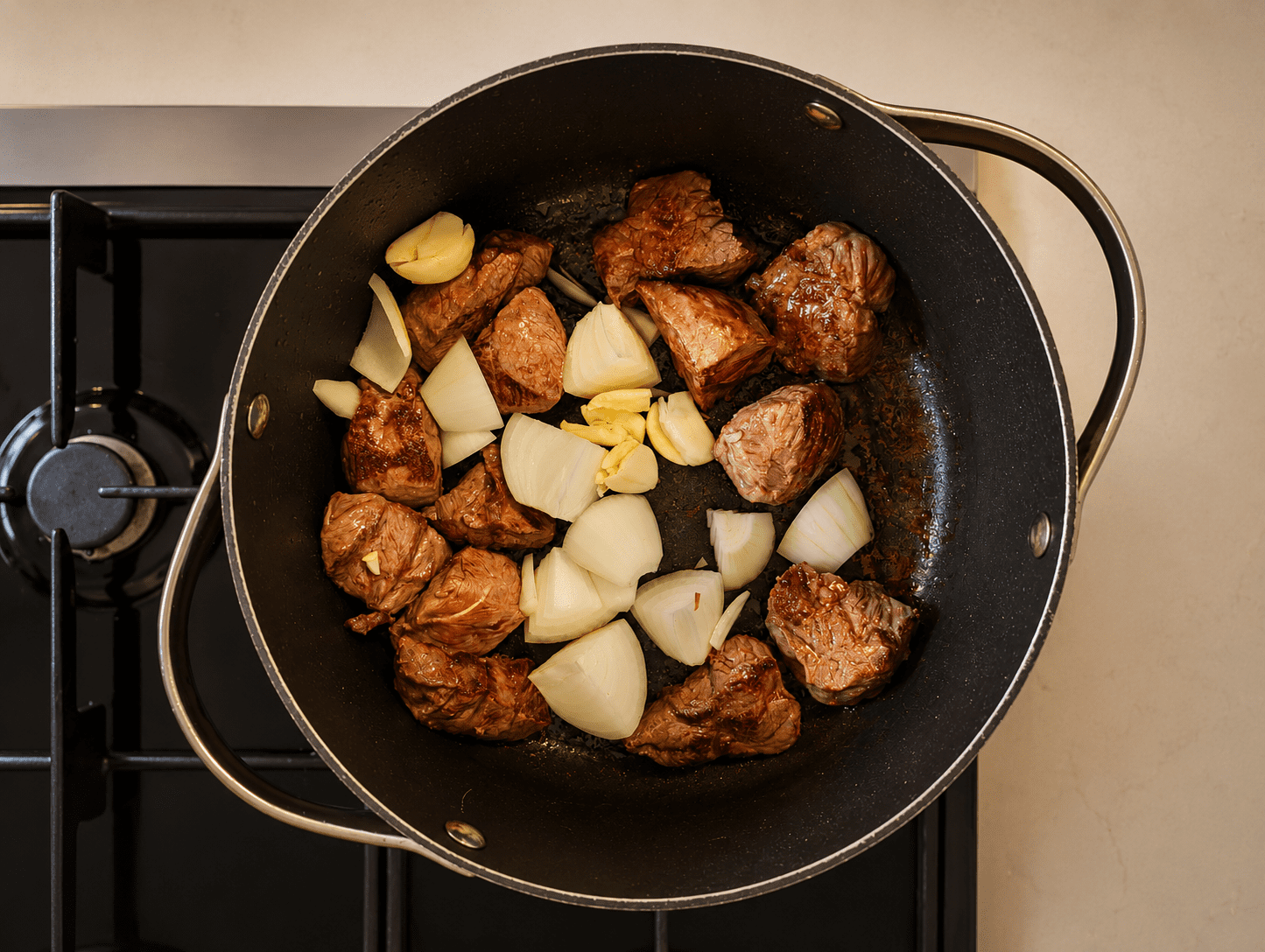 Beef cubes browning on high heat with chunks of onion and garlic in a black pot