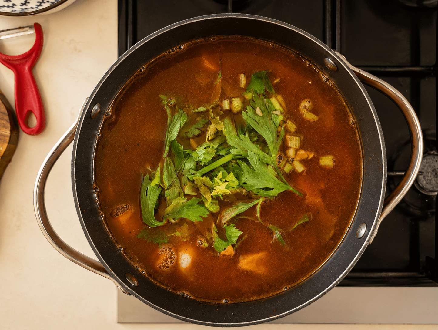 A full pot of red goulash broth with celery leaves on top settling into a long low simmer