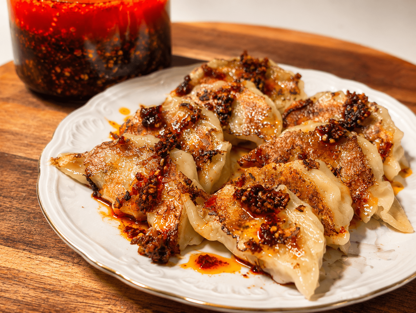 Pan-fried Japanese gyoza dumplings on a white plate generously drizzled with homemade chili oil, jar of chili oil in the background
