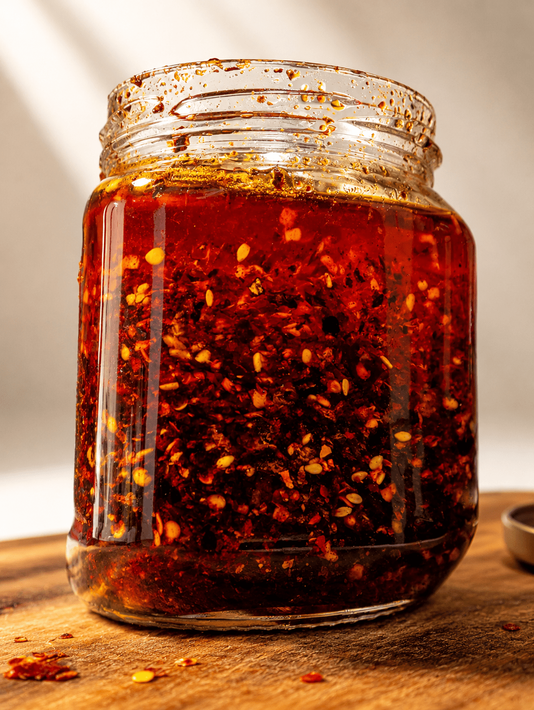 Glass jar of homemade chili oil on a wooden board, side-lit, glowing deep red with visible chili flakes and sesame seeds suspended in the oil
