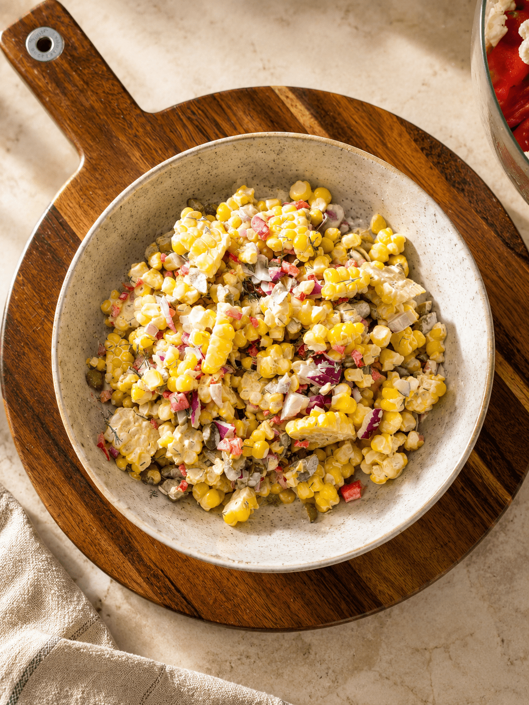 Top-down bowl of creamy corn salad on a wooden board, bright yellow corn kernels mixed with finely diced red bell pepper, red onion, pickles and fresh dill
