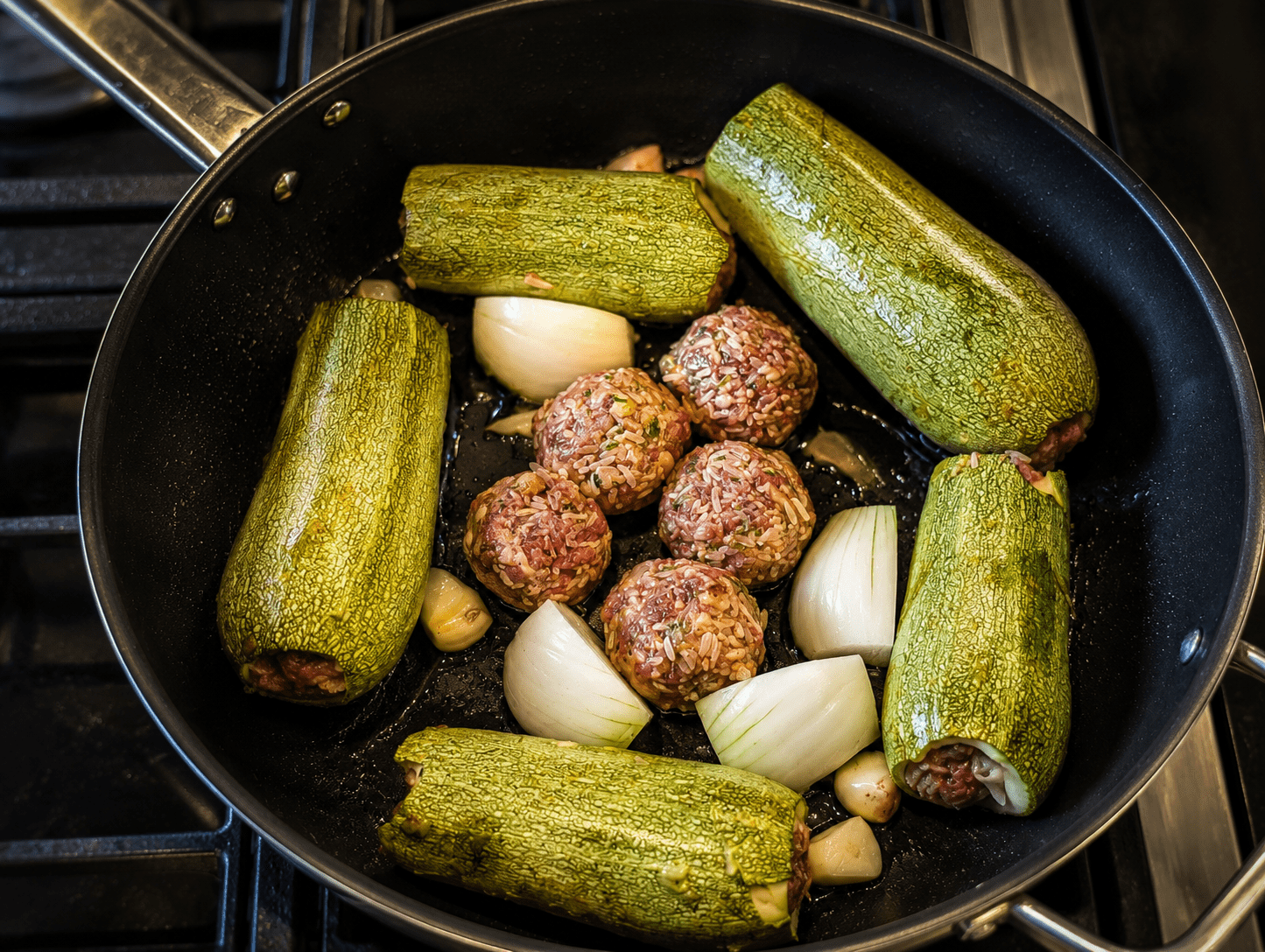 Stuffed zucchini and meatballs searing in a pot with quartered onion and halved garlic cloves