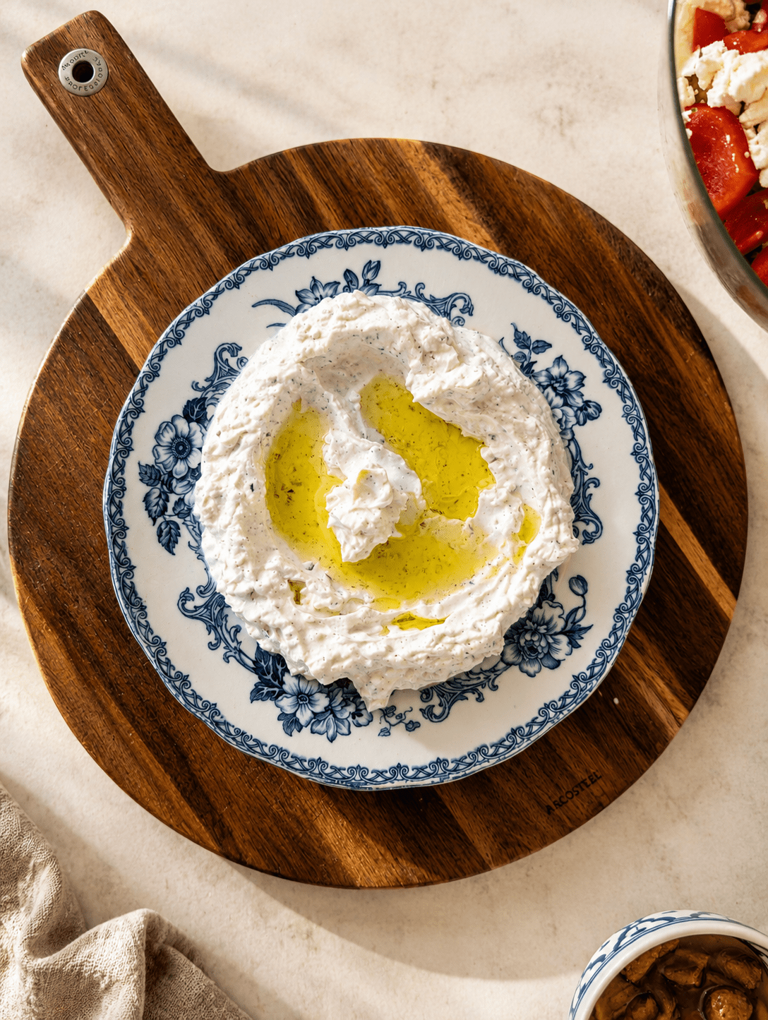 Homemade tzatziki served on a blue floral plate on a wooden board, thick and creamy with grated cucumber and dill mixed in, a generous well of olive oil pooled in the center