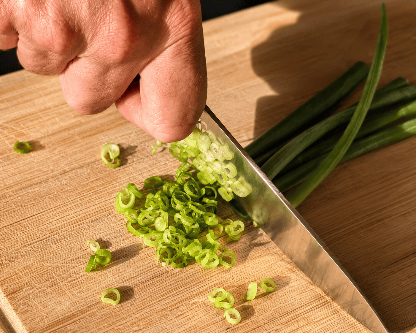 Green onions being thinly sliced into rings on a wooden cutting board with a chef knife