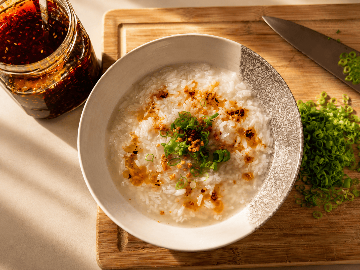 A bowl of congee topped with chili oil, crispy garlic and green onions, next to a jar of homemade chili oil