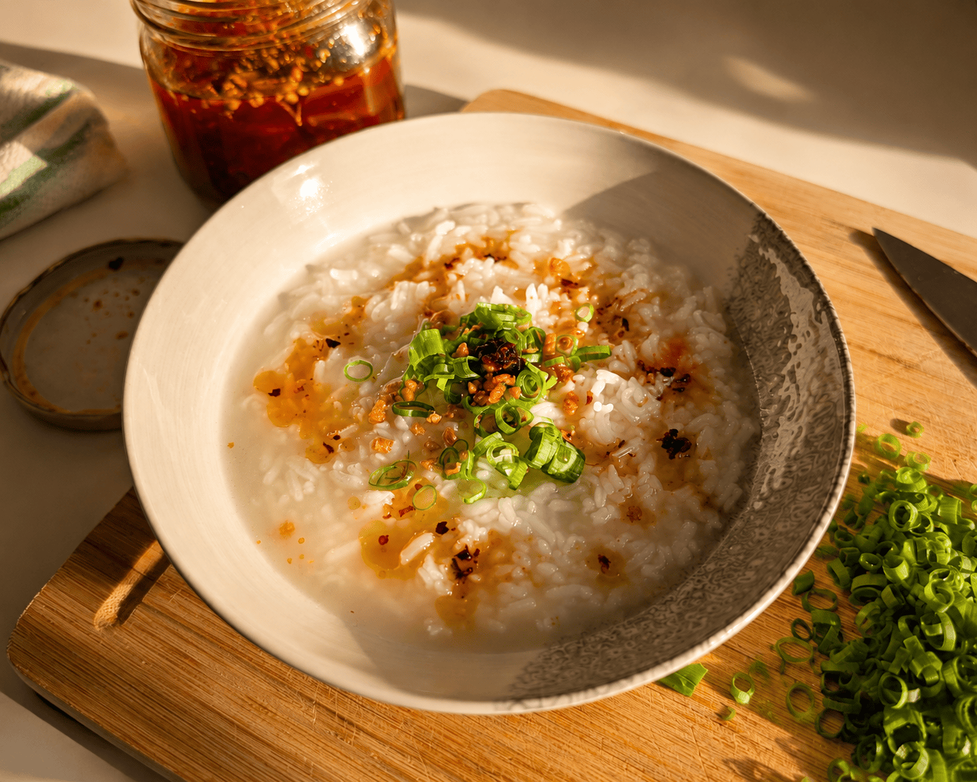 A close up bowl of creamy congee with chili oil pooling on the surface, crispy garlic and sliced green onions on top