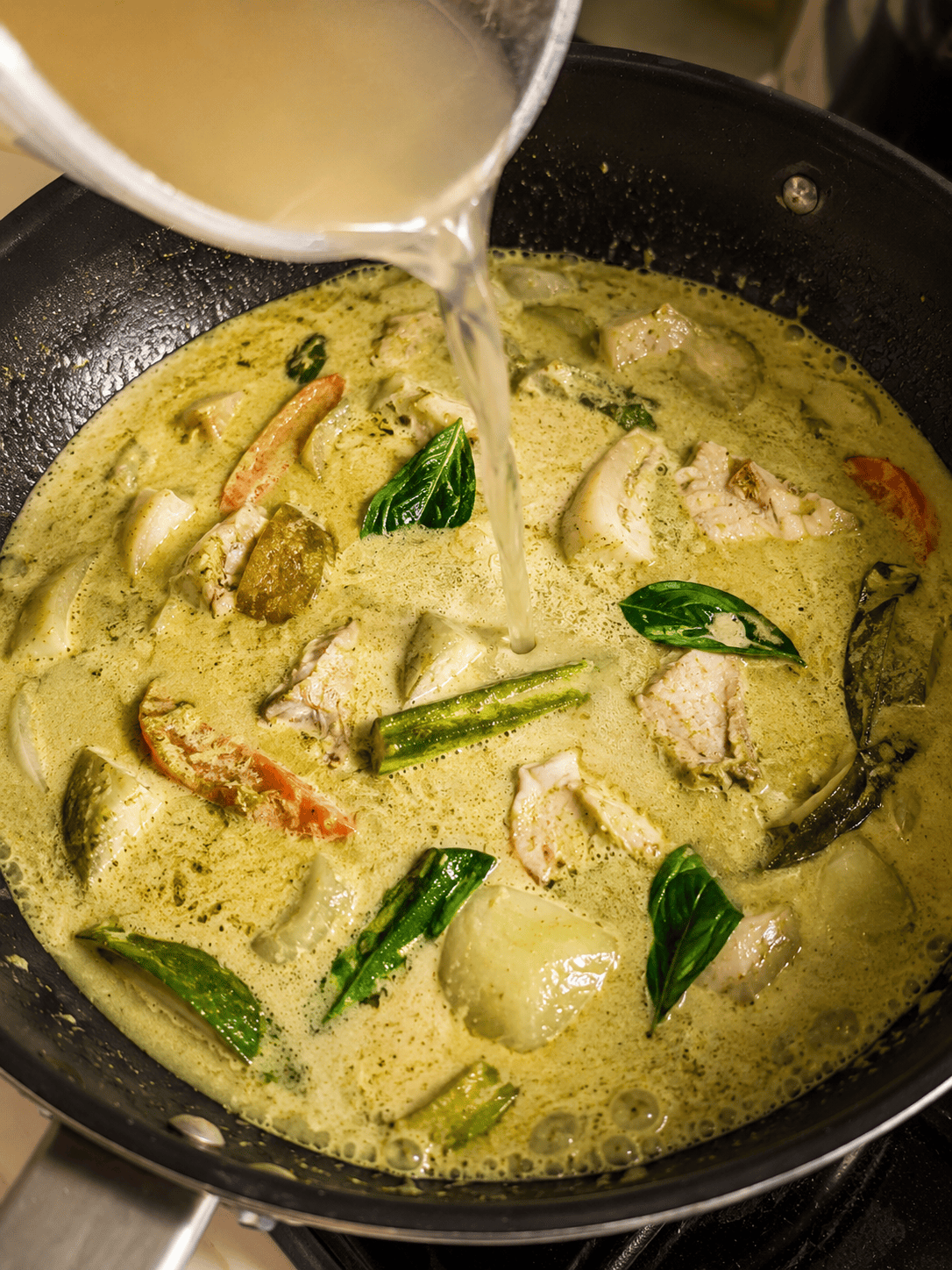 Chicken stock being poured into a pan of Thai green curry with fish and basil leaves