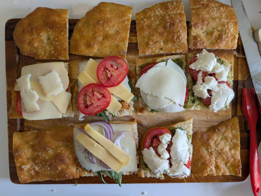 Close side-on cross-section of a focaccia sandwich showing manchego, red onion, and arugula layers next to another sandwich with mozzarella and tomato