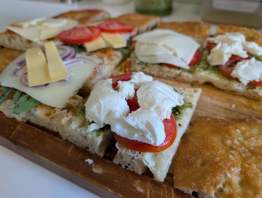 Angled close-up of open-face focaccia sandwiches topped with manchego, tomato, red onion, mozzarella, and pesto
