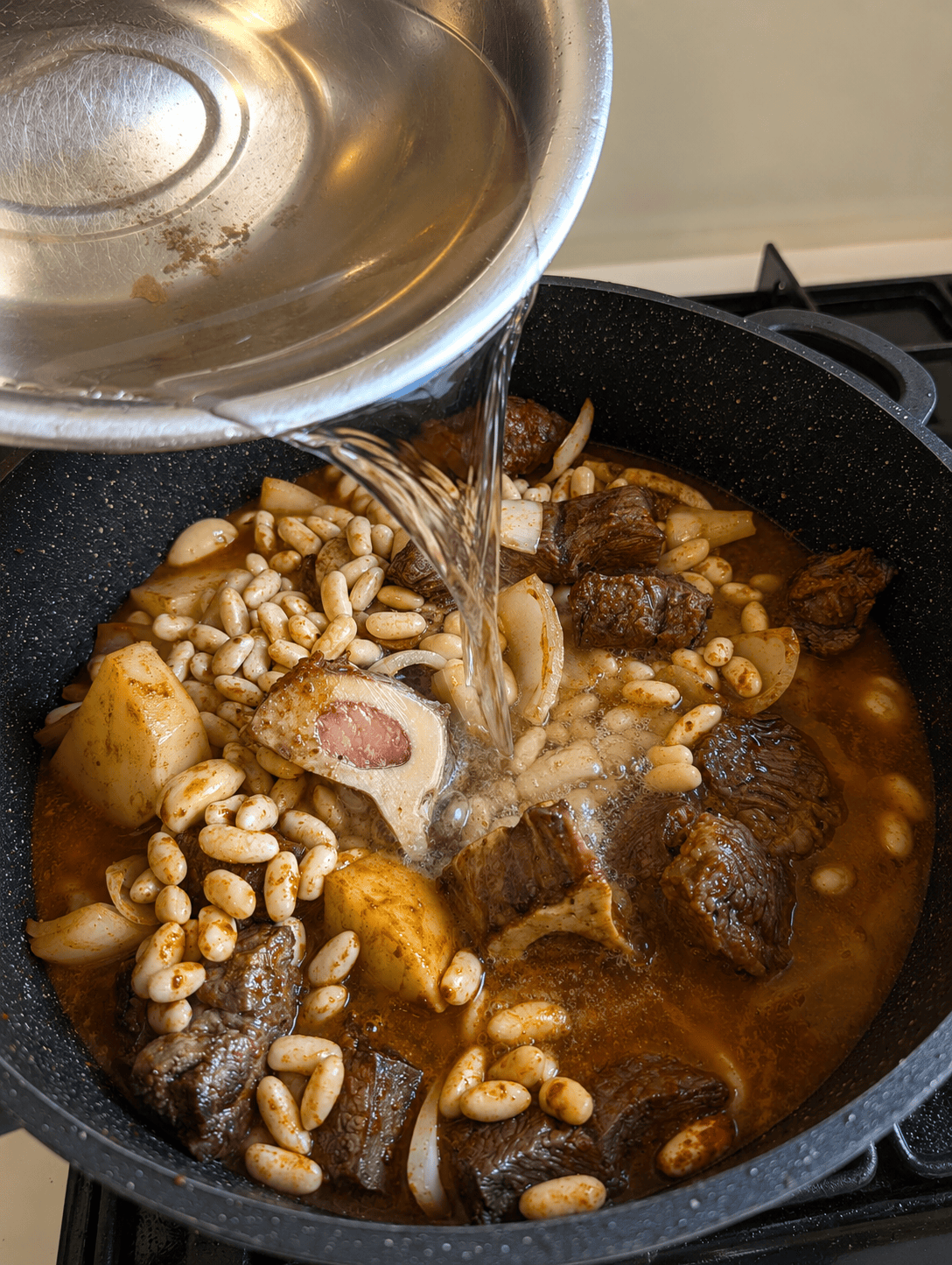 Water being poured into a pot of beef, white beans, potato, and onion coated in paprika