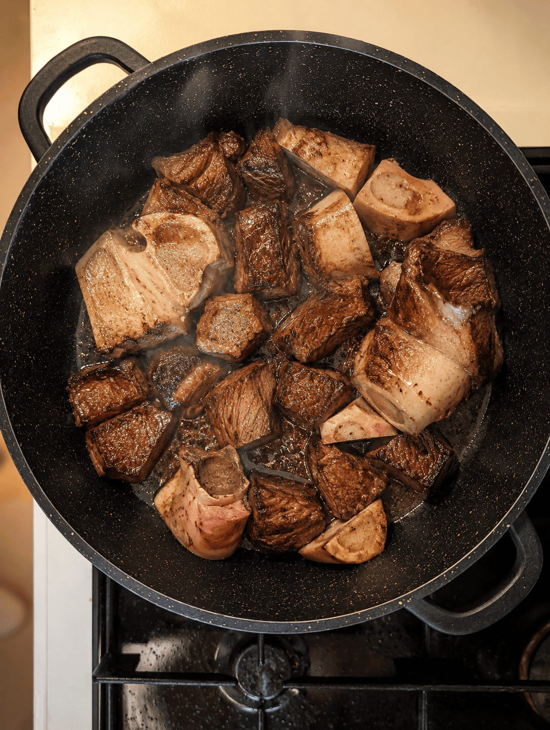 Beef cubes and marrow bones searing and browning in a large pot with oil, steam rising