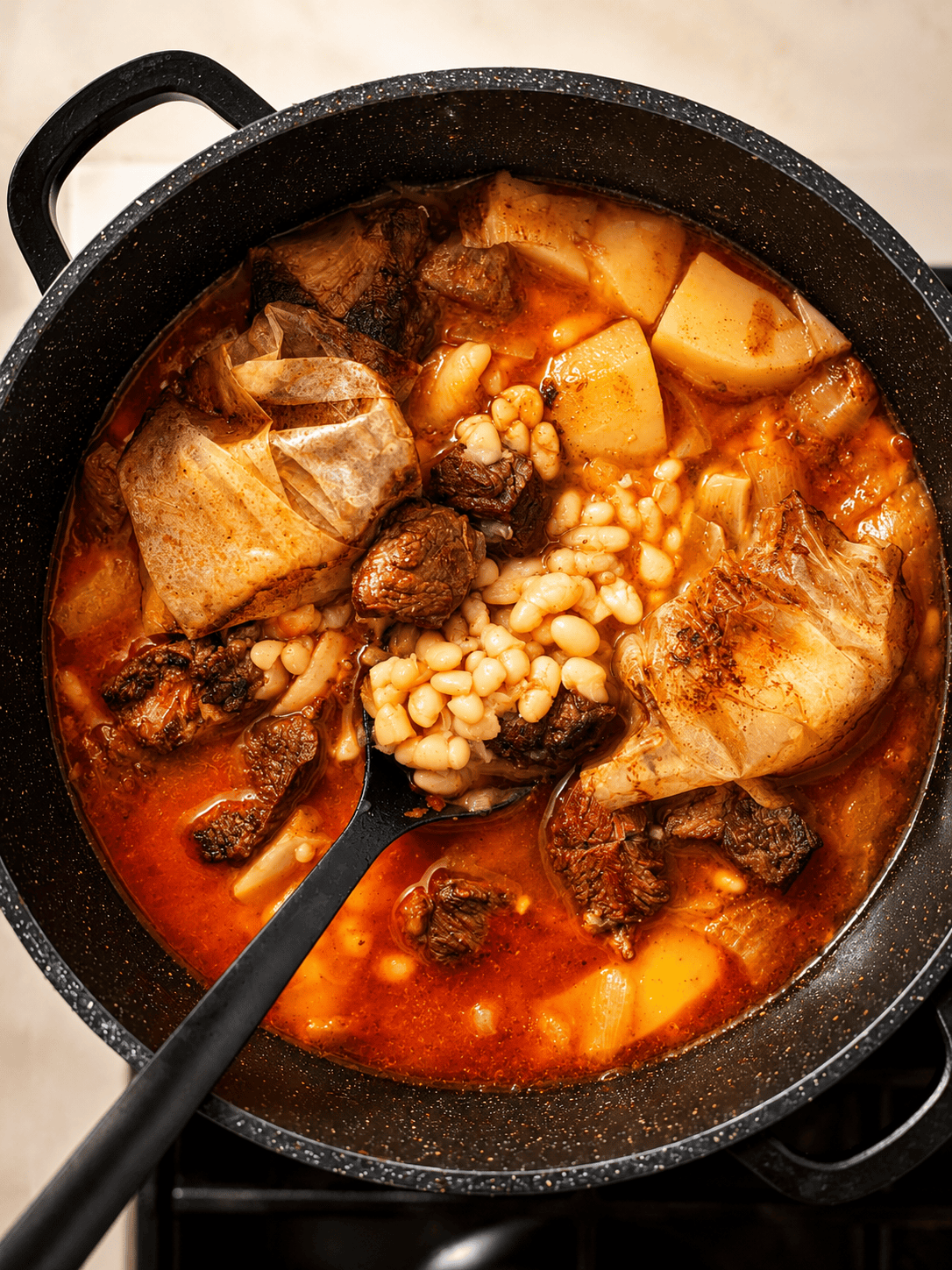 Spoon lifting tender beef, white beans, and a parchment-wrapped egg out of the finished tbicha stew
