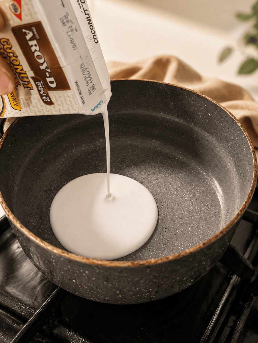 Coconut cream being poured from a carton into a small dark saucepan on the stove to make the sauce for mango sticky rice