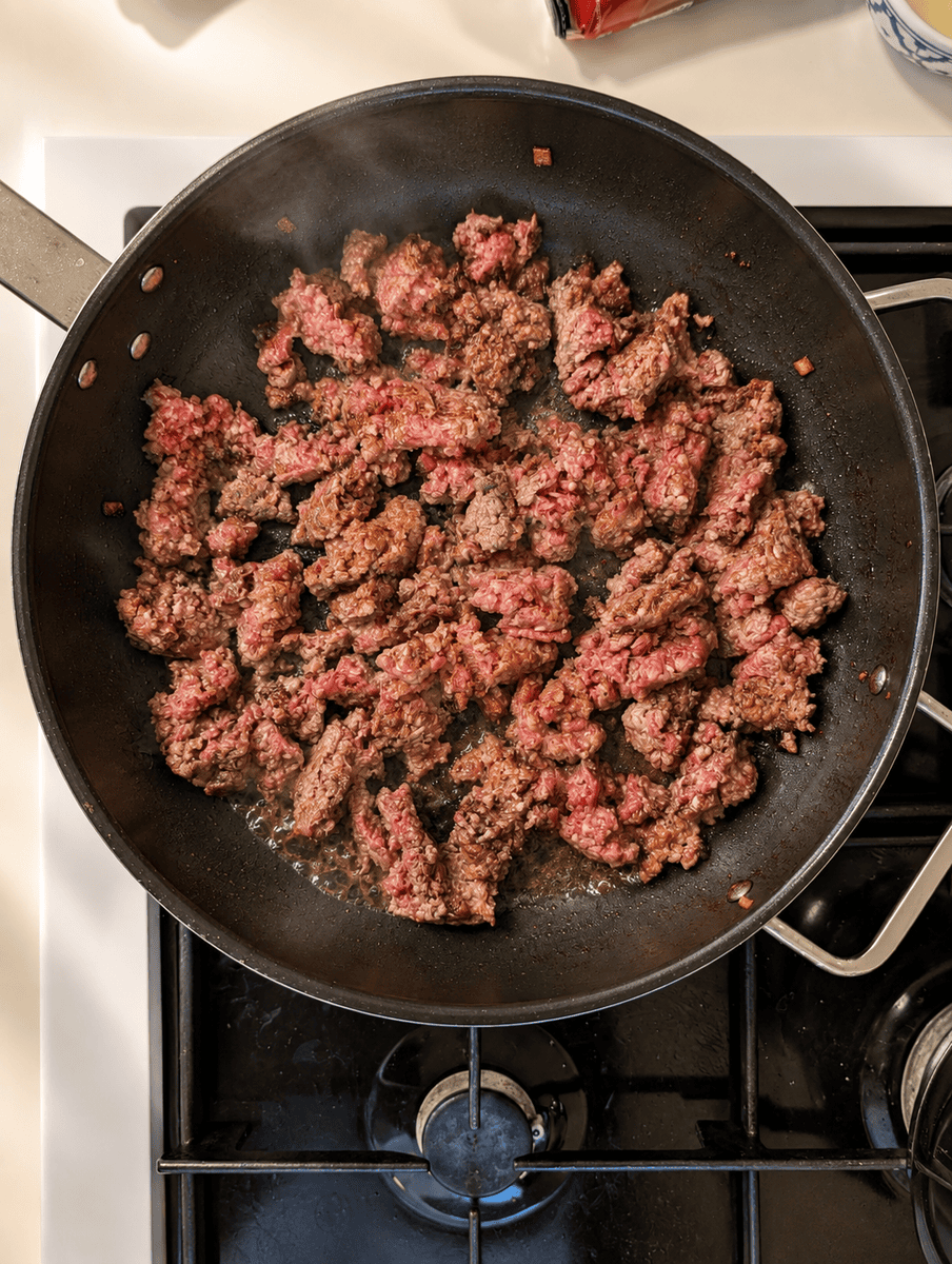 Ground beef breaking up and beginning to brown in a wide black frying pan on the stove