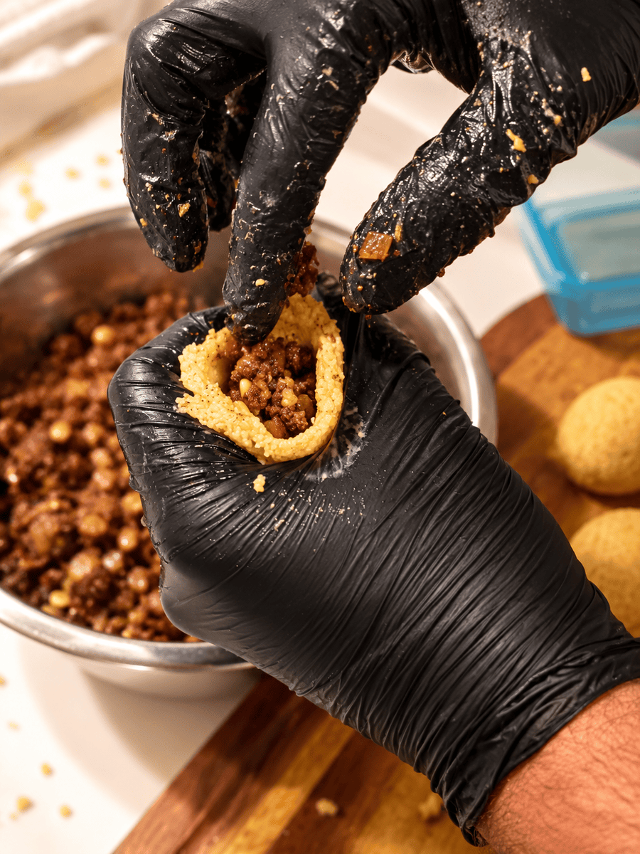 Gloved hands packing spiced beef and pine nut filling into a hollow bulgur shell before sealing it closed