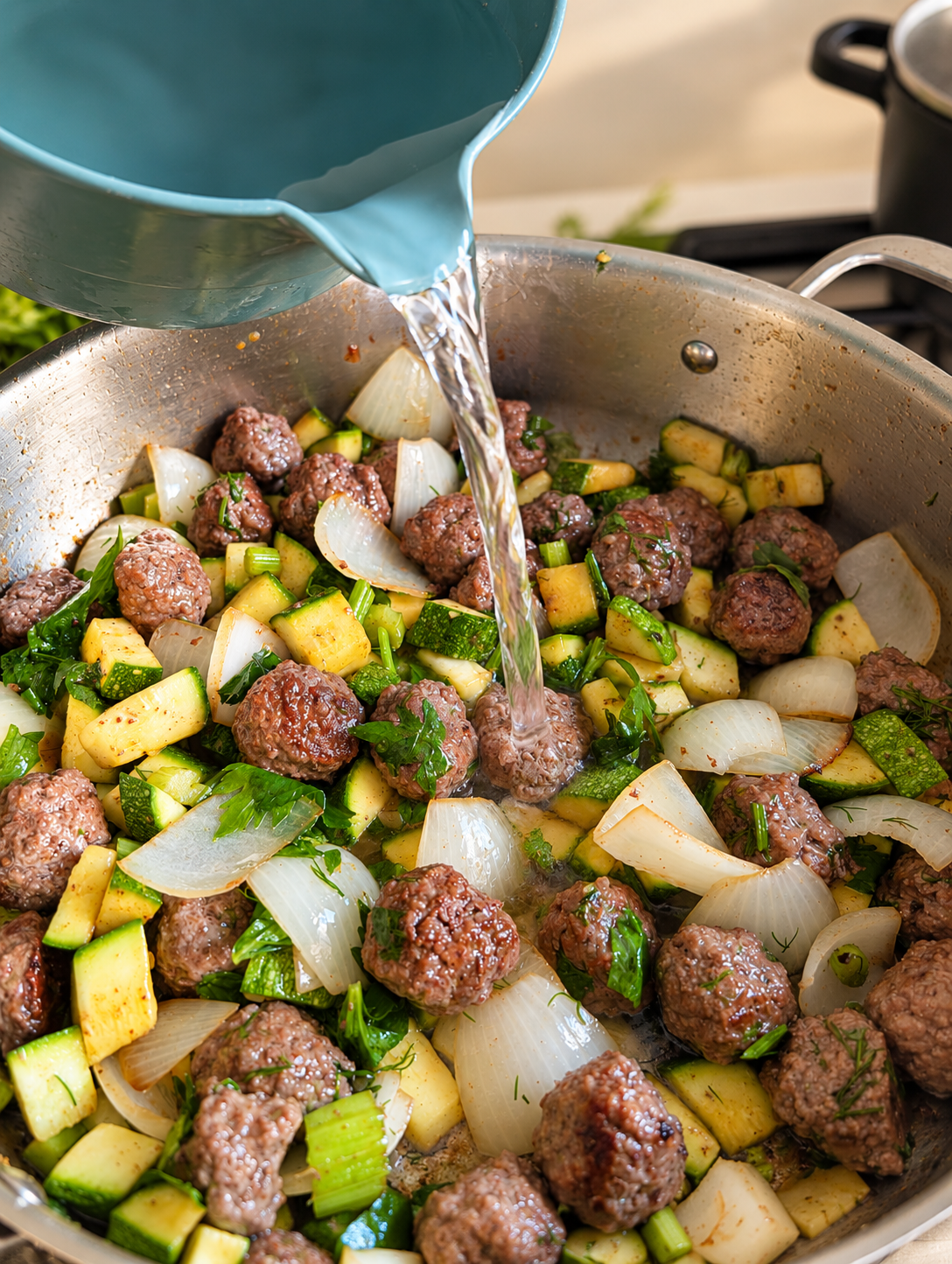 Water being poured from a blue measuring jug into a pan of meatballs, zucchini and herbs