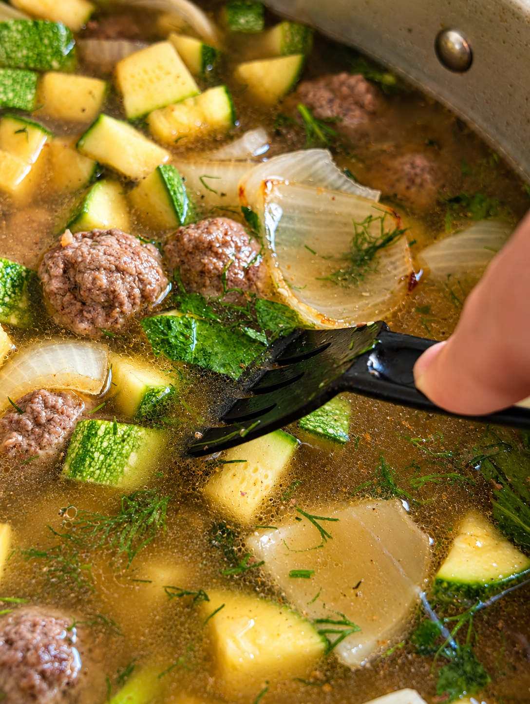 Close-up of meatball zucchini soup simmering in a pot with a slotted spatula moving meatballs and zucchini through the broth