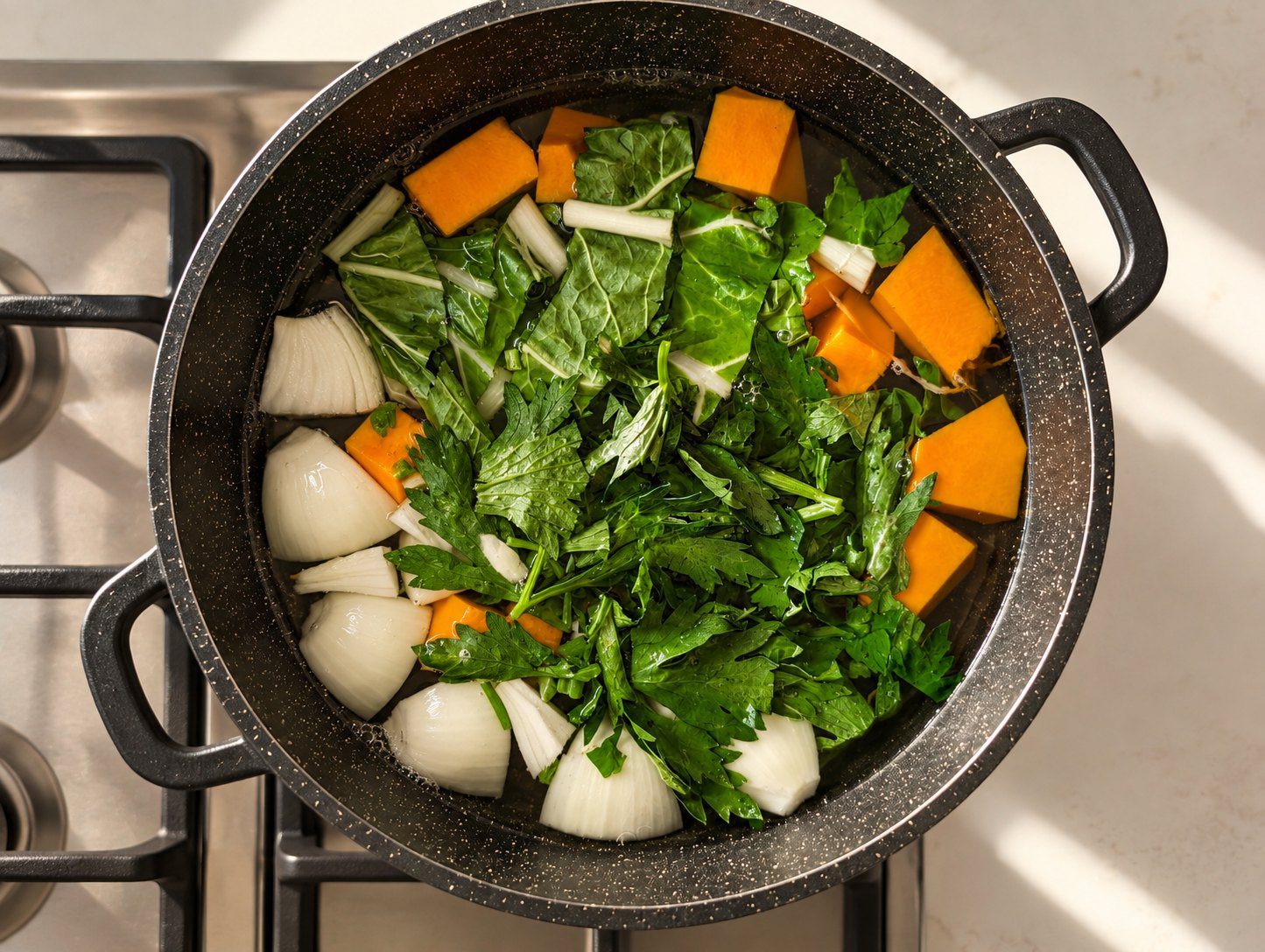 Overhead view of a black pot filled with water, chunks of pumpkin, onion quarters and green leaves before cooking